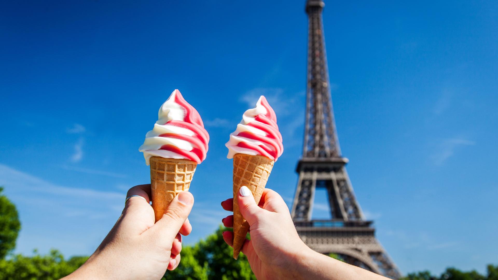 Two hands holding up pink and white swirled ice cream cones in front of the Eiffel Tower Paris