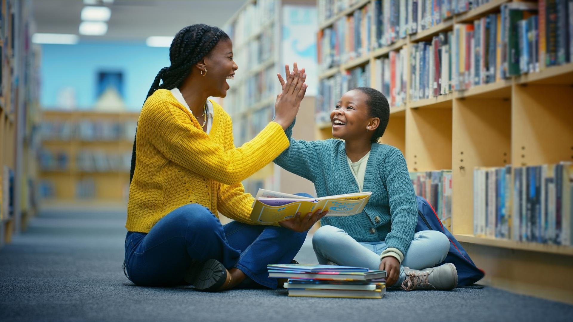 Mother and daughter high fiving sitting on the floor in a library with a stack of picture books in front of them
