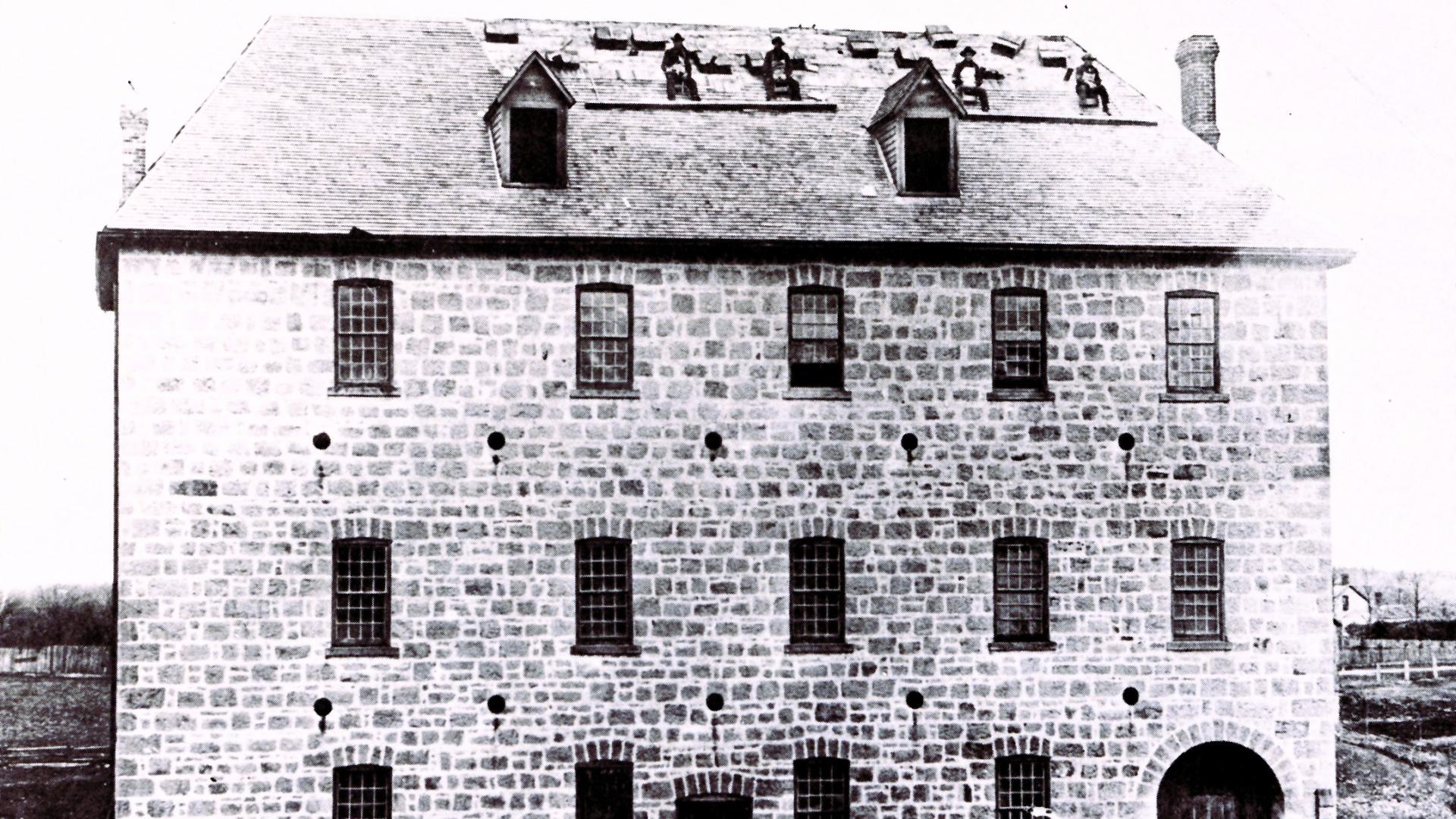 Three story nineteenth century stone mill with several workers sitting on the peaked roof having a break