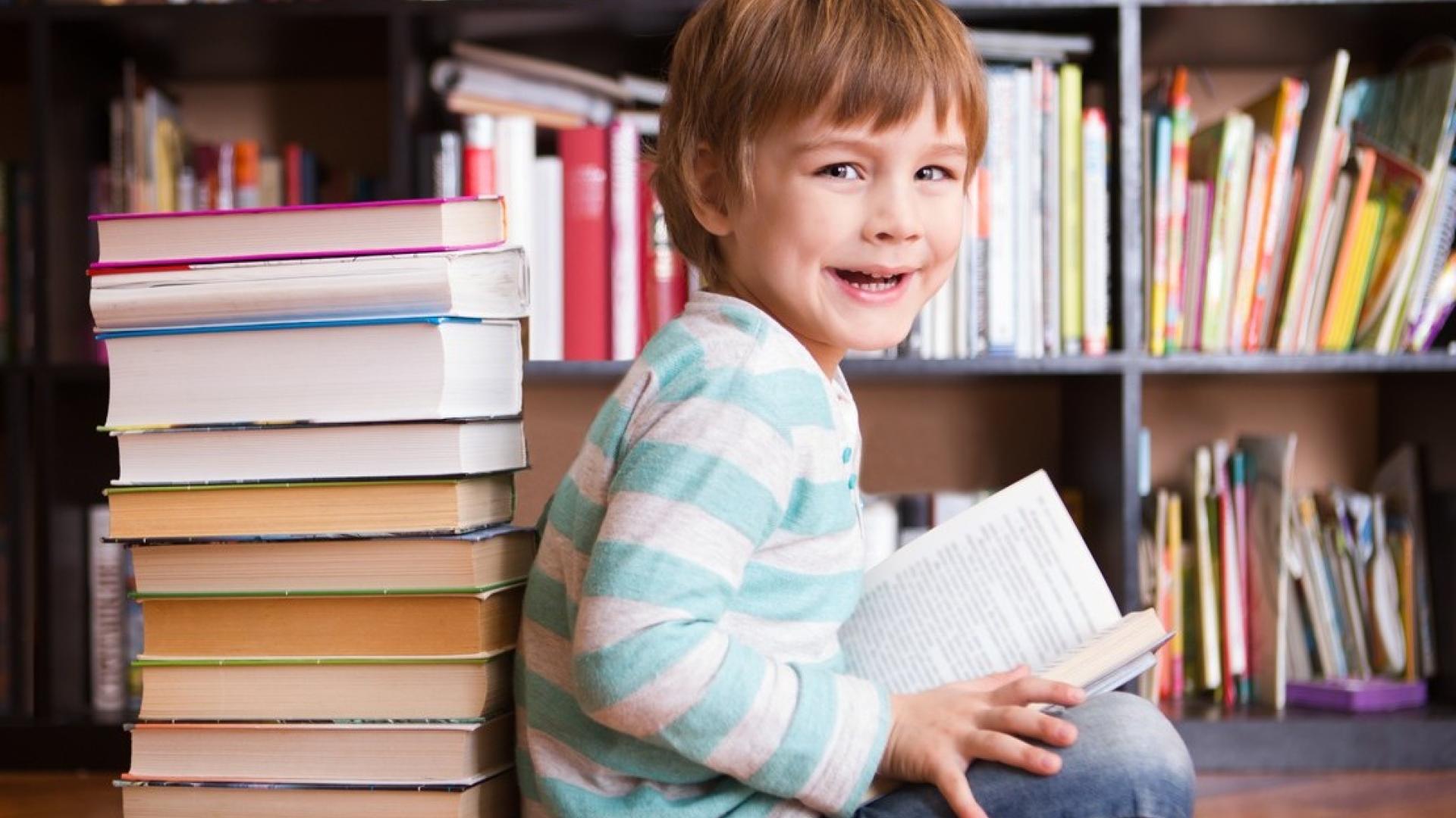A little boy smiles reading a book in front of a bookcase