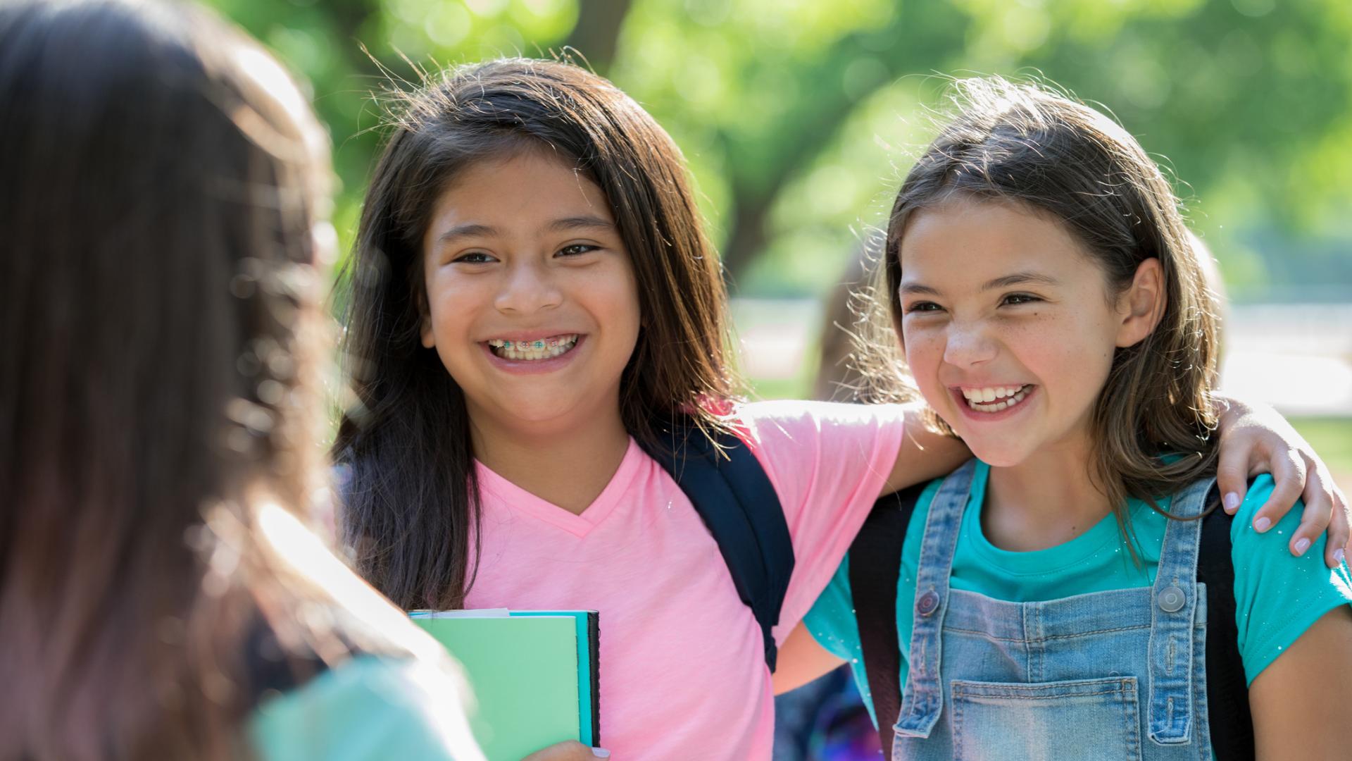 Tween girls laugh together wearing backpacks and holding notebooks