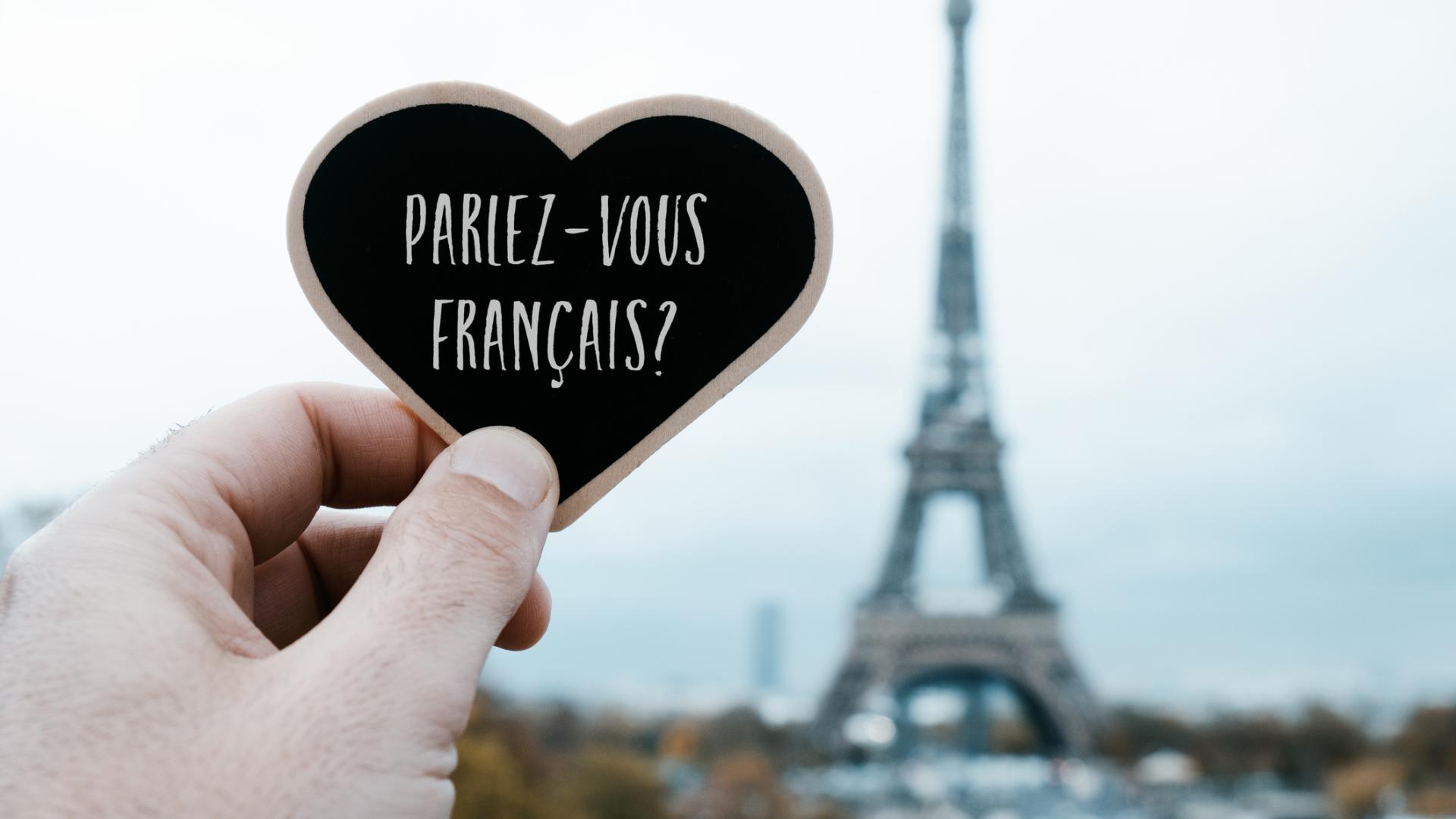 A hand holds up a heart that reads Parlez Vous Francais in front of the Eiffel Tower in Paris