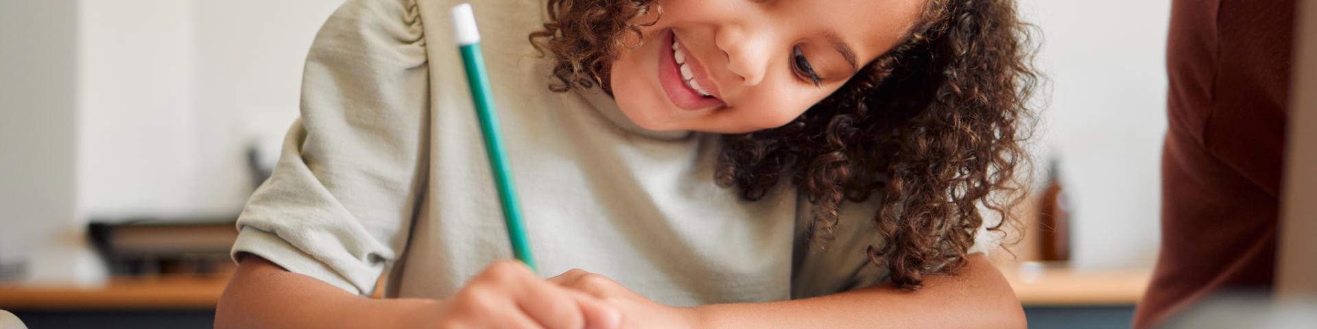 Smiling little girl with curly black hair writing in a notebook with a teal blue pencil
