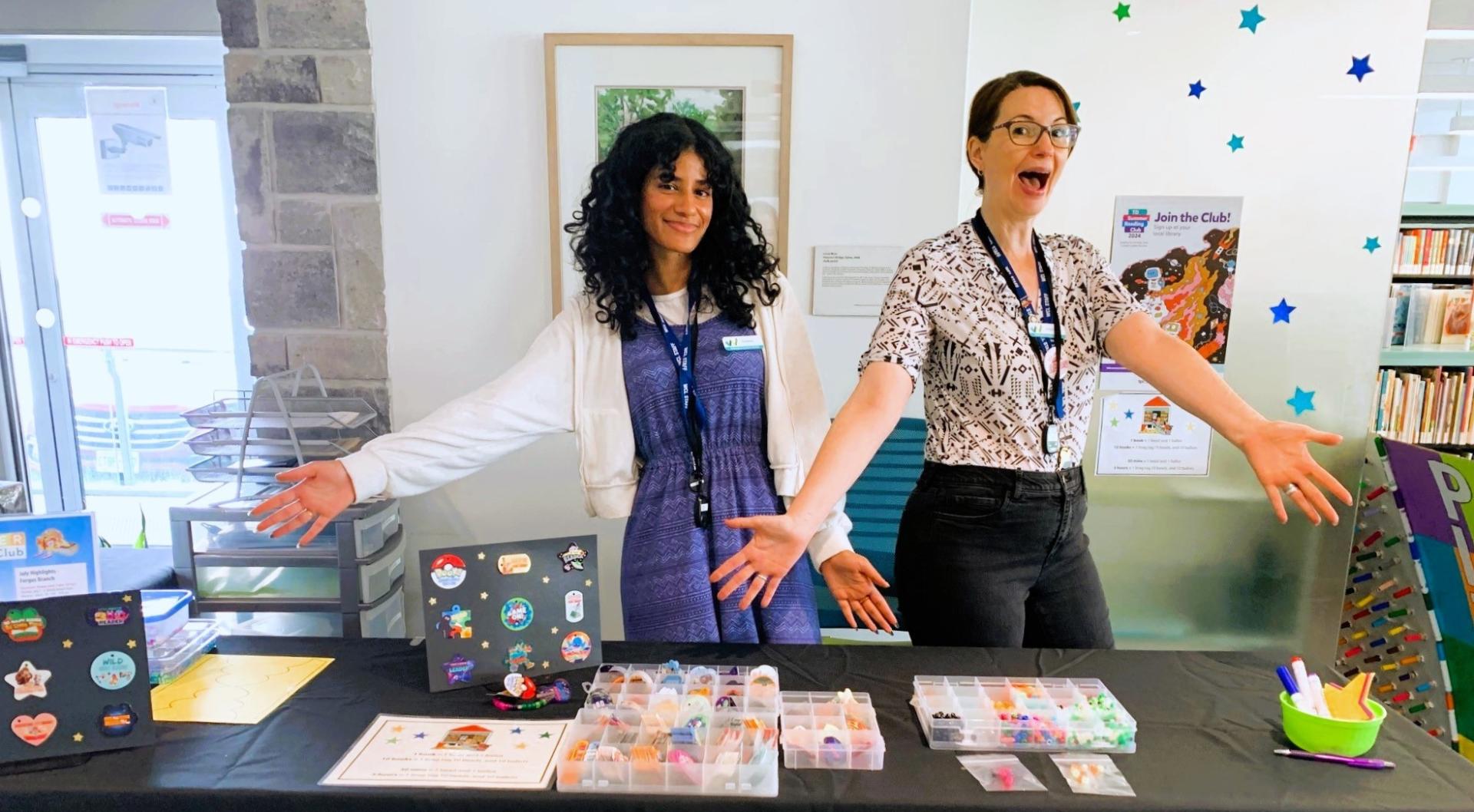 Two women stand behind table with beads and brag tags displayed for summer reading