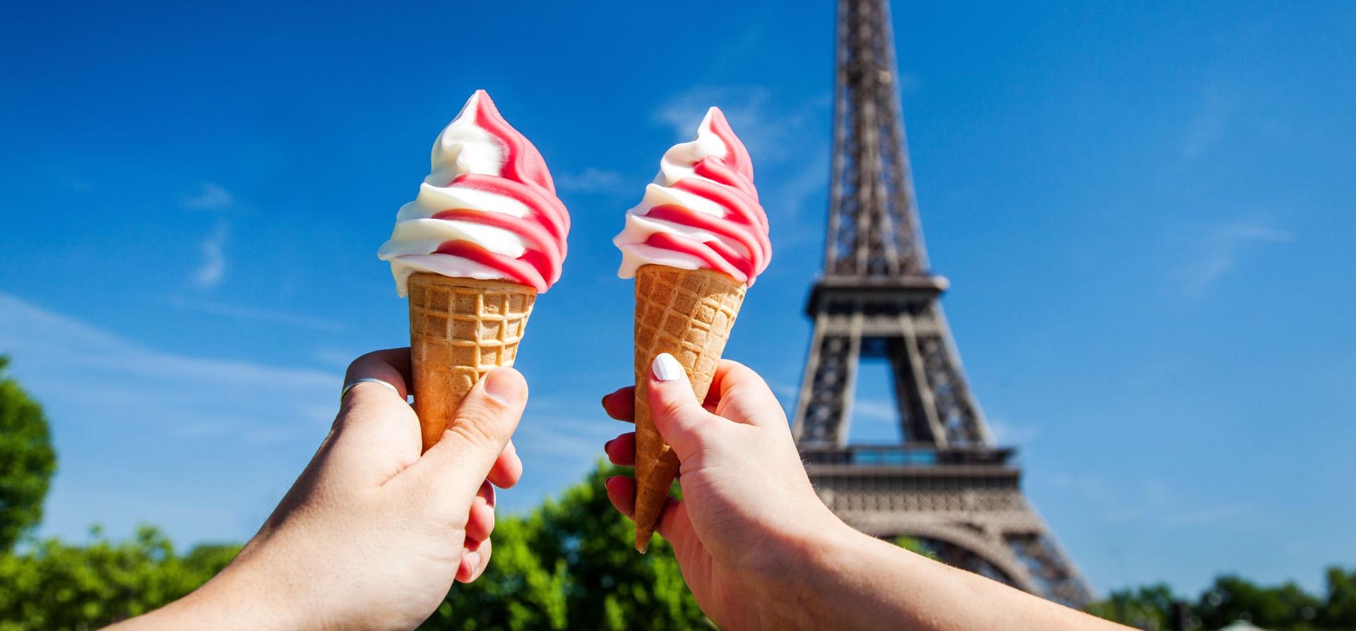 Two hands holding up pink and white swirled ice cream cones in front of the Eiffel Tower Paris