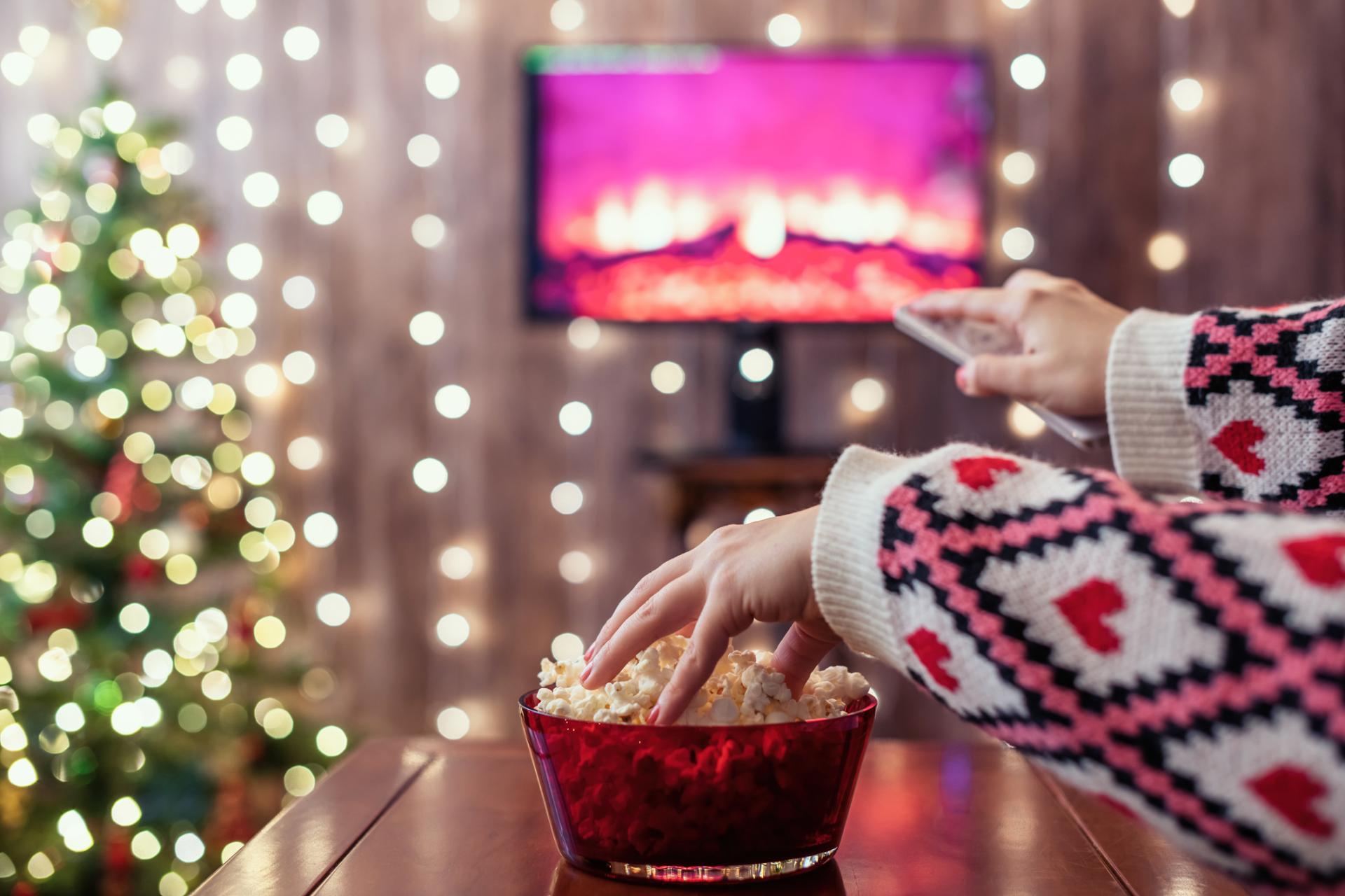 One hand in the popcorn bowl and one hand holding a remote pointing at a TV screen with a fireplace displayed on it