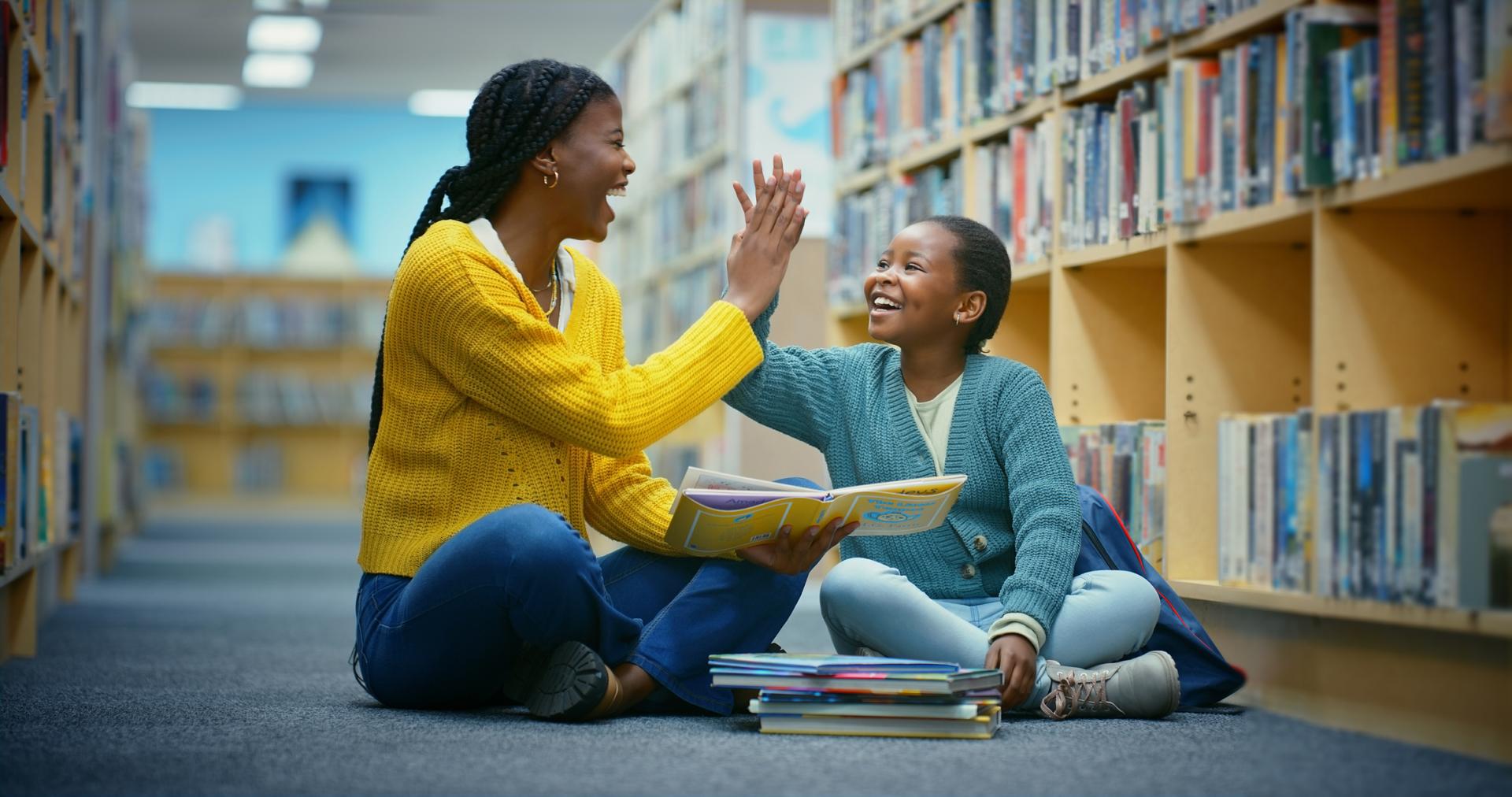 Mother and daughter high fiving sitting on the floor in a library with a stack of picture books in front of them