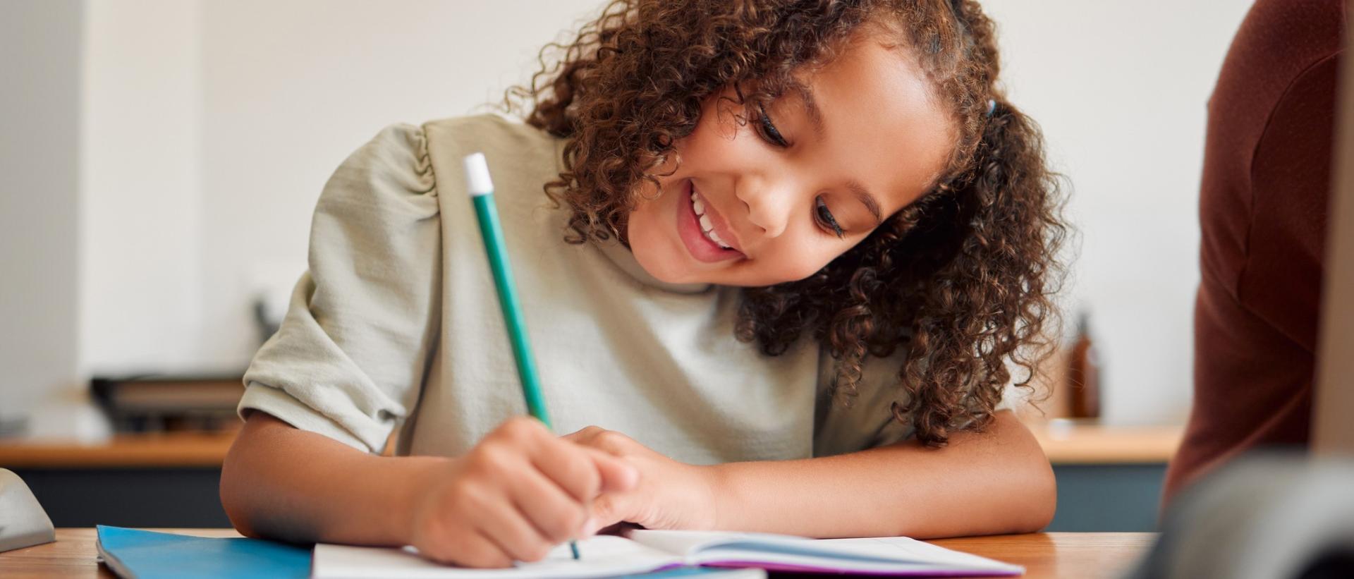 Smiling little girl with curly black hair writing in a notebook with a teal blue pencil